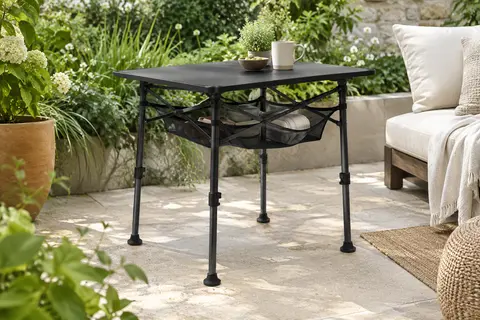 Table pliante noire Anbte à trois hauteurs avec filet sous le plateau, installée sur une terrasse végétalisée.