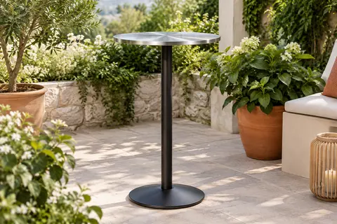 Table haute noire Casaria avec plateau rond en inox et piètement réglable, installée sur une terrasse végétalisée.