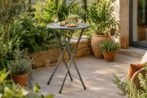 Table haute ronde grise à pieds métalliques pliables installée sur une terrasse végétalisée