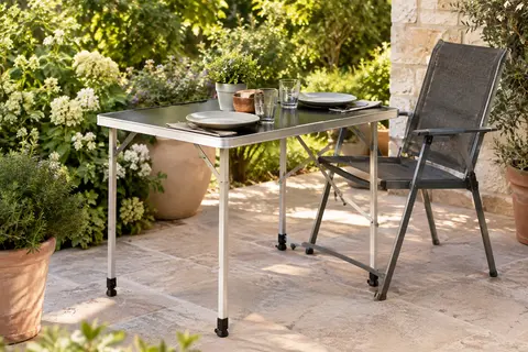 Table pliante de camping à plateau noir en MDF et pieds en aluminium, installée sur une terrasse végétalisée.