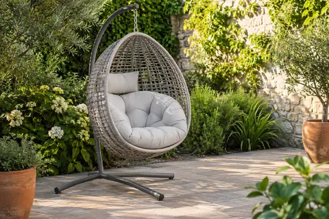 Fauteuil suspendu gris clair à effet rotin avec coussin et support métallique sur une terrasse végétalisée.