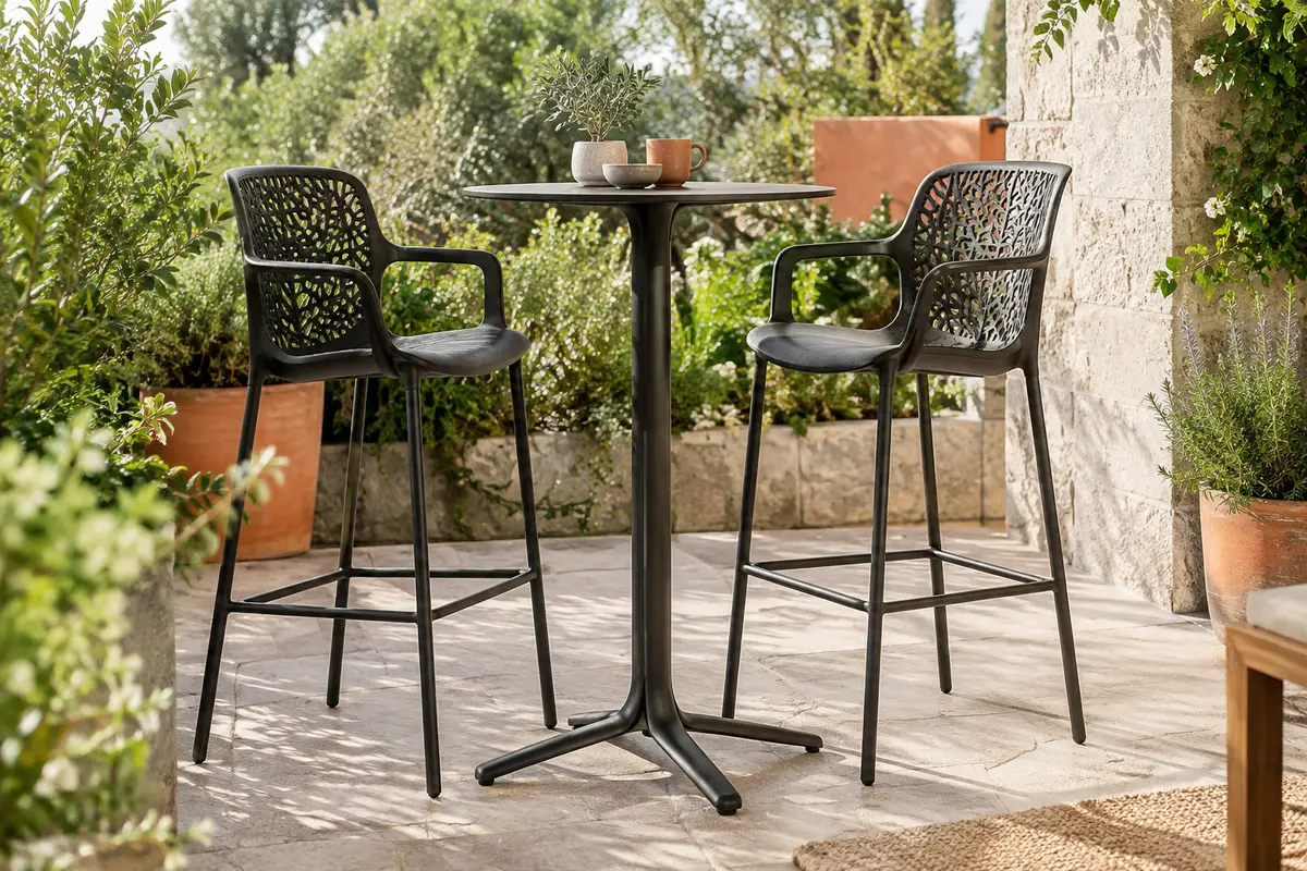 Deux tabourets de bar noirs à dossier ajouré installés autour d’une table haute sur une terrasse végétalisée.