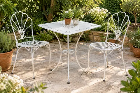 Ensemble bistrot blanc en métal avec motifs floraux, table carrée et deux chaises sur une terrasse végétalisée.